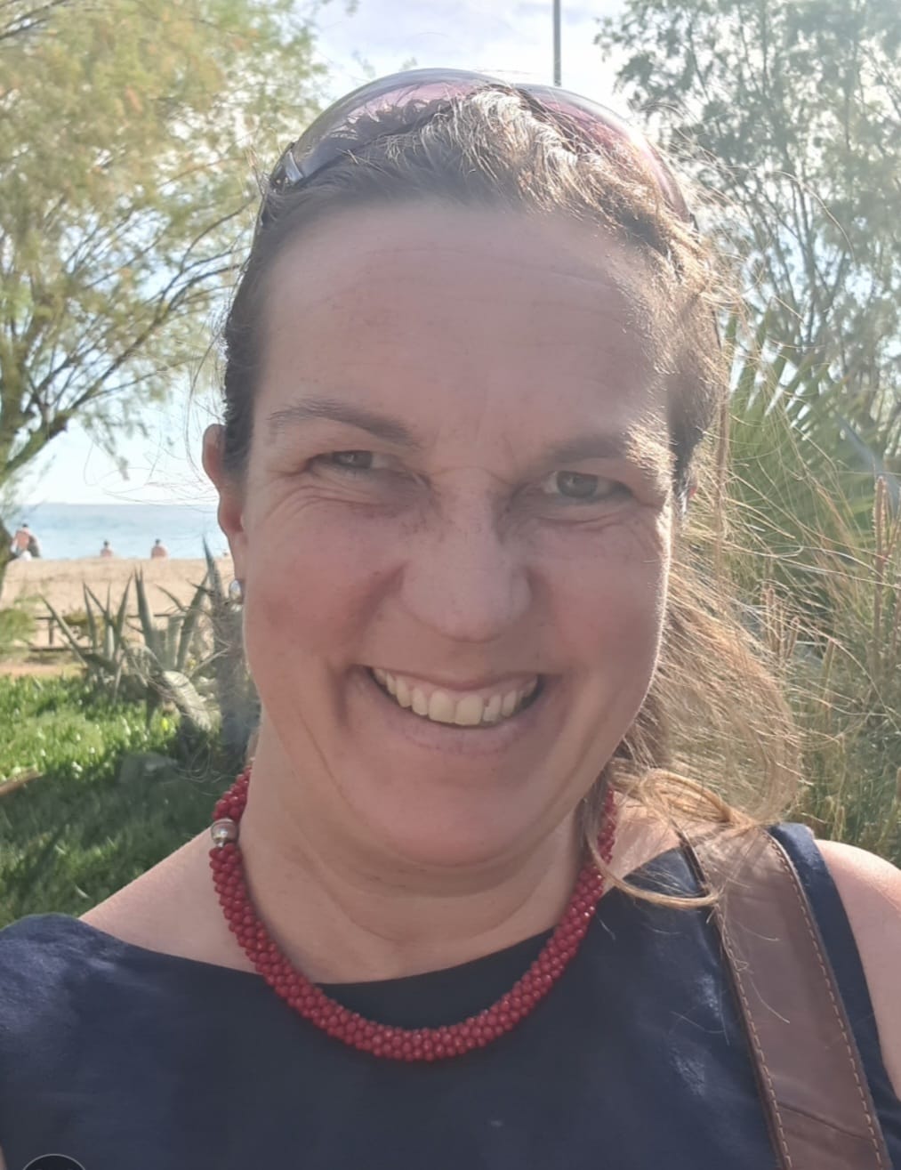 002 konstanze Baur Auf dem Bild sieht man Konstanze Baur im Profil. Sie lacht fröhlich in die Kamera. Ihre brünetten Haare hat sie nach hinten gebunden. Sie trägt eine rote Kette und ein dunkelblaues Oberteil. Auf ihrer linken Schulter sieht man den Riemen einer Tasche. Im Hintergrund sieht man Natur, Strand und ein Stück vom Meer. Das Wetter ist sehr schön und die Sonne scheint.